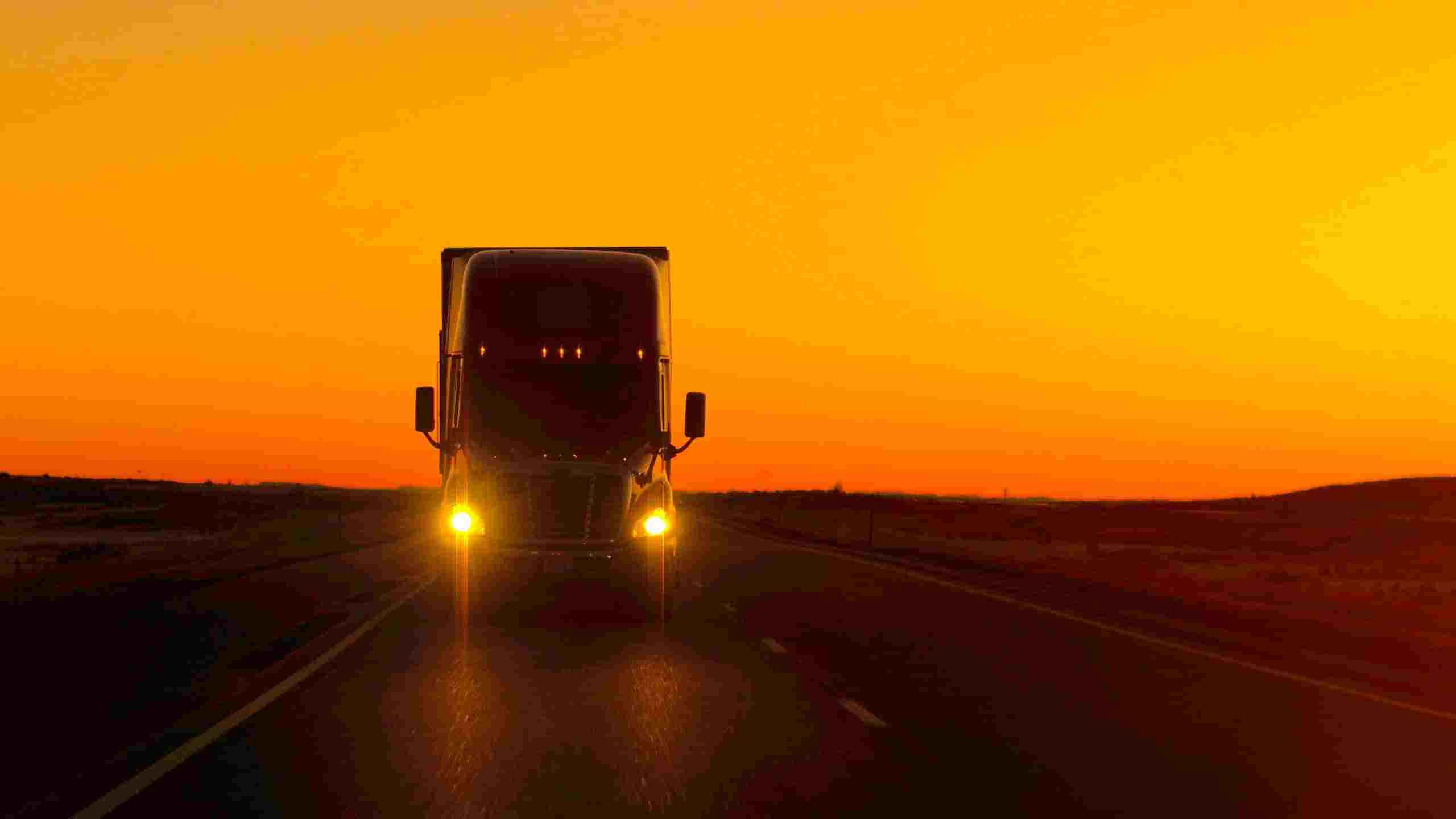 Big rig driving towards the camera with its lights with a sunrise in the background.