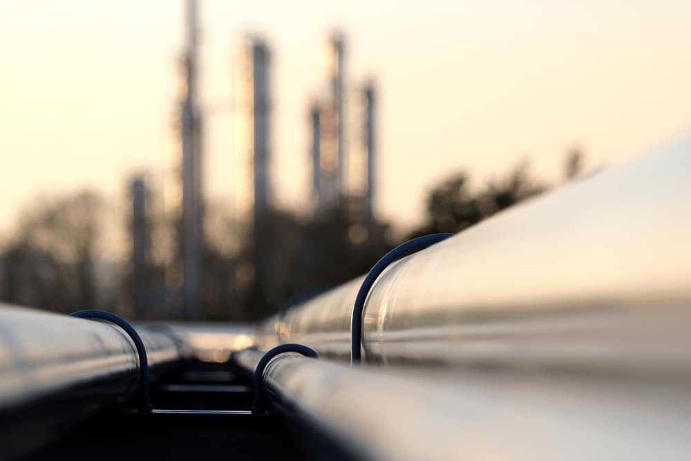 Closeup of pipelines with a refinery in the background.