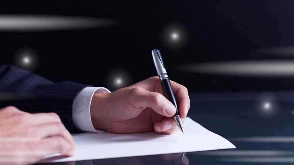 Man holding a fancy pen writing on paper on a desk.