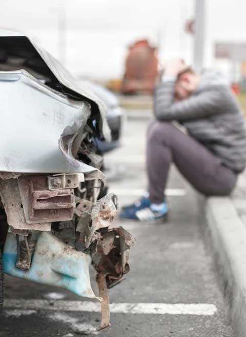 A distraught woman sitting on the curb next to a wrecked car.