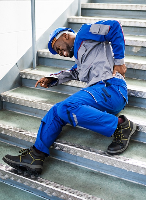 A man in a blue work uniform holding his back in pain after falling on the stairs.